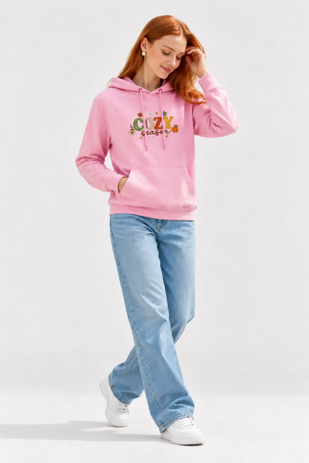 Woman with red hair wearing a pink custom embroidered hoodie with “COZY season” autumn embroidery, wide-leg light blue jeans and white sneakers