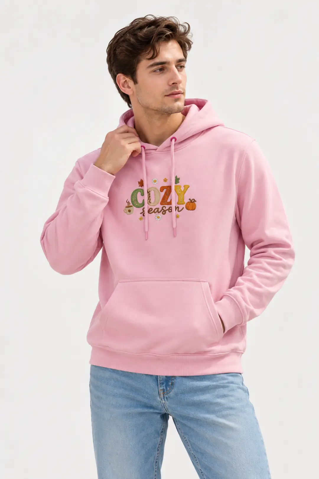 Man wearing a pink custom embroidered hoodie with “COZY season” text, pumpkin, mug and daisy embroidery on the chest, paired with light blue jeans