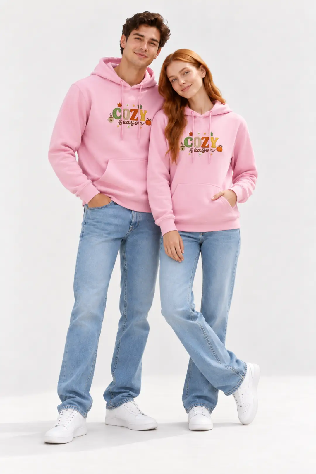 Man and woman posing together in matching pink custom embroidered hoodies with colorful “COZY season” autumn embroidery, styled with light blue jeans and white sneakers