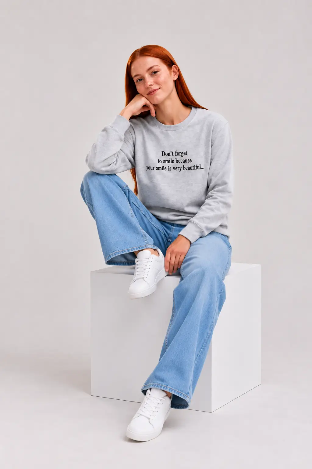 Woman sitting in grey unisex sweatshirt with black embroidered smile slogan and light-wash wide-leg jeans