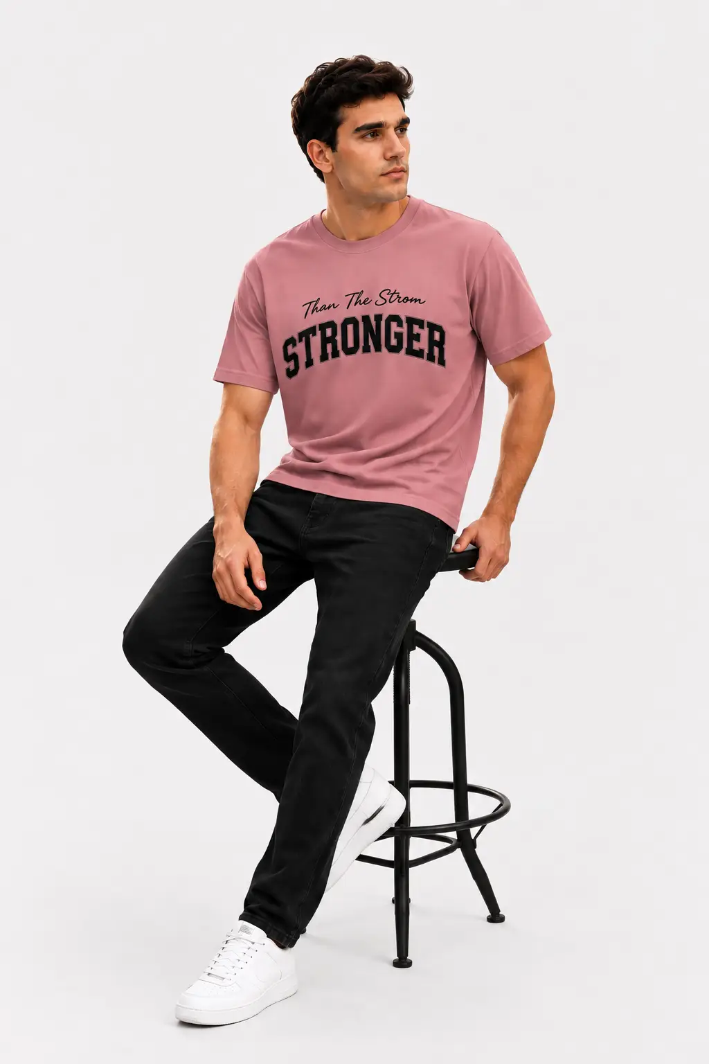 Man seated on a black stool wearing a dusty rose oversized t-shirt with black “STRONGER” print, black jeans and white sneakers