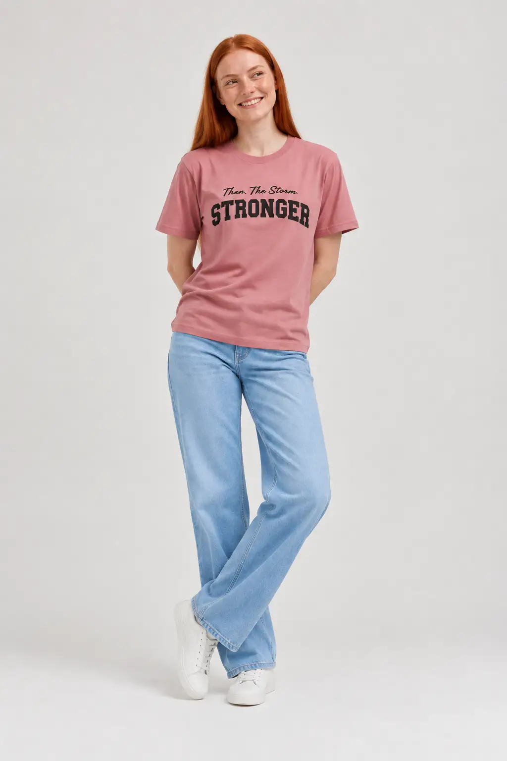 Smiling woman with red hair wearing a dusty rose t-shirt with black “STRONGER” print and wide-leg light blue jeans